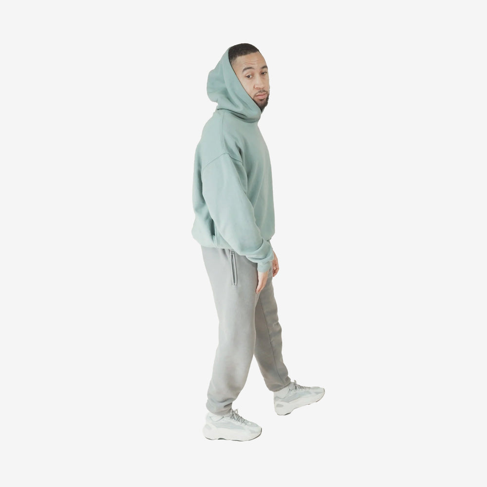 Man wearing Aqua Round Hoodie with hood up, paired with gray pants and white sneakers.