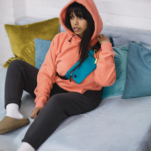 female model sat on aqua suede sofa with multi-coloured pillows, wearing a teal shoulder bag, cozy peach hoodie with the hood up, core gray premium jogging pants