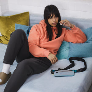 female model sat on aqua coloured sofa, wearing tailored jogging pants in core gray colour, cozy peach hoodie, blue bag and multi-coloured cushions