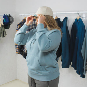 female model wearing hat, azure blue cotton hoodie, ash joggers, multi-coloured clothes hung up in background