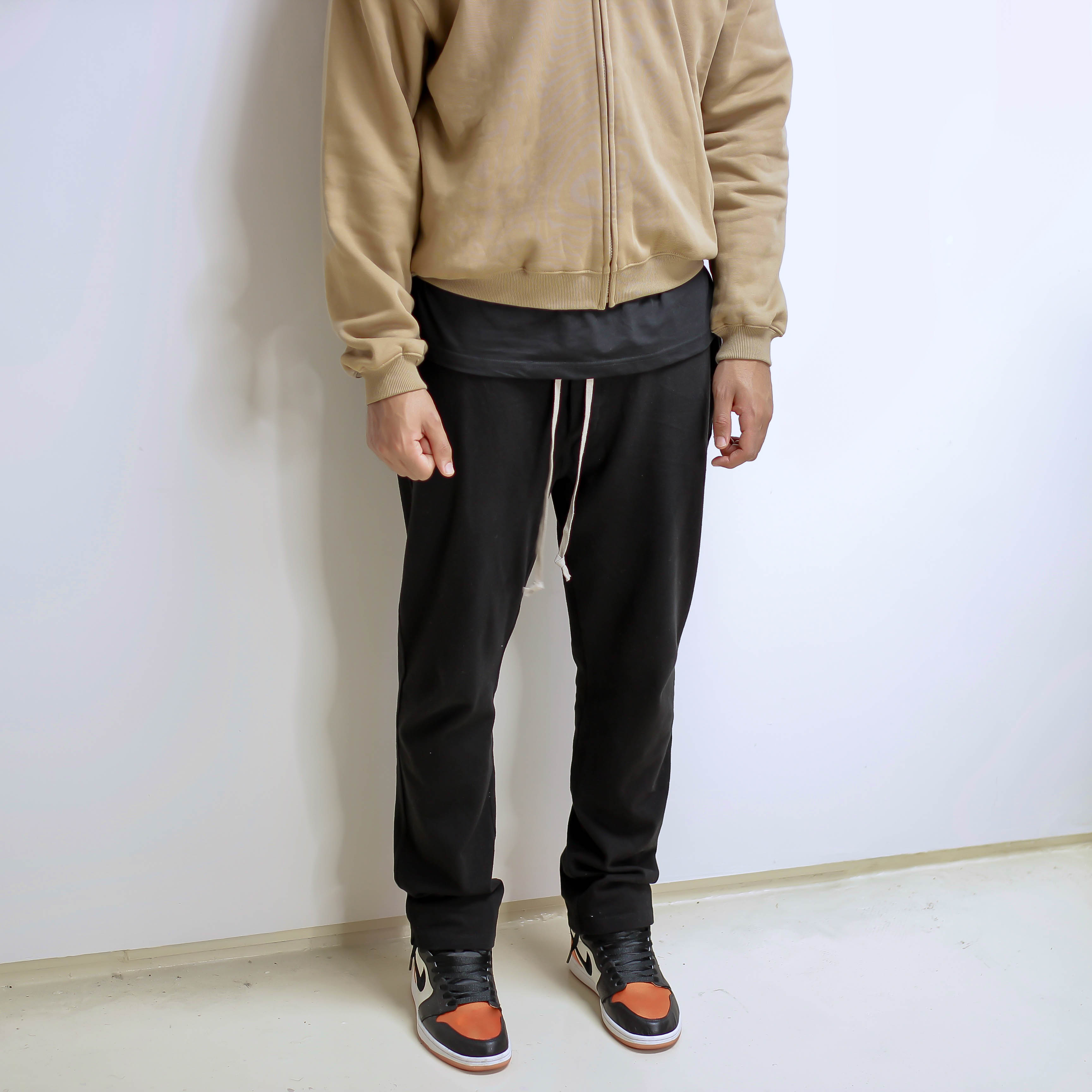 Black drawstring pants worn with sneakers and a beige jacket against a white background.